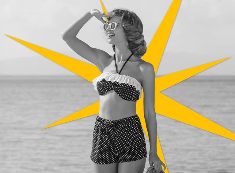 Woman wearing beach attire, standing on a rock by the ocean.