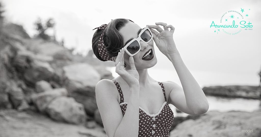 Woman at the beach in 1950's swimsuit attire and holding sunglasses on her face. (Model)